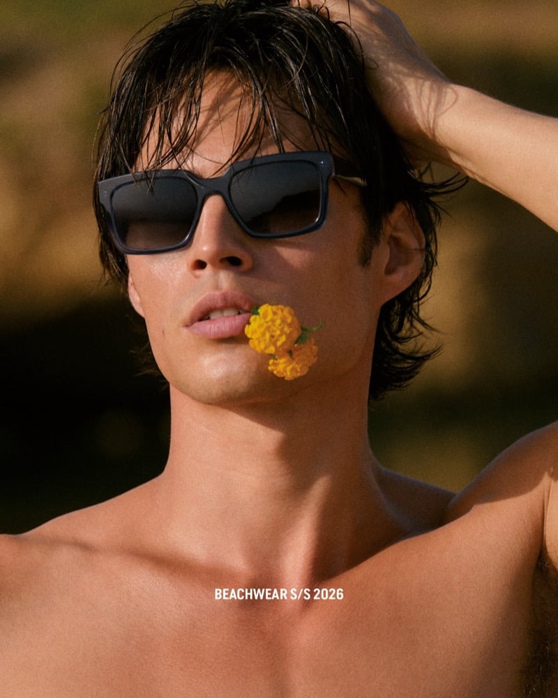 Close-up of Louis in square black acetate sunglasses holding a yellow marigold flower between his lips against a golden coastal backdrop