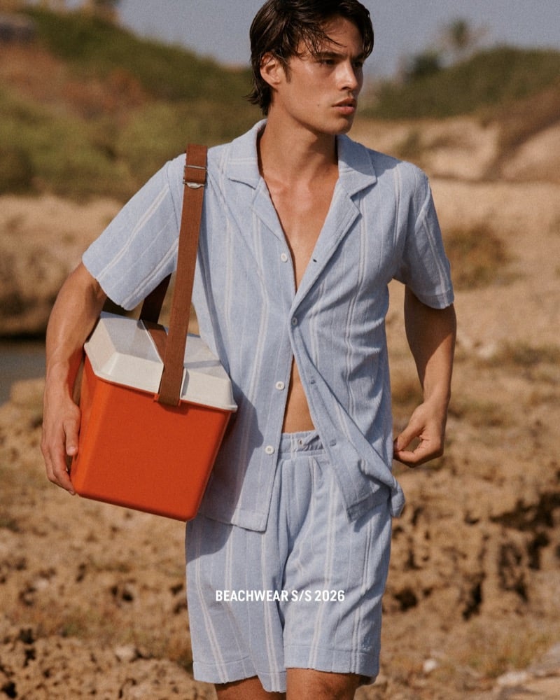 Louis walks across sandy ground in a pale blue terry cloth camp-collar shirt and matching striped shorts, carrying an orange cooler bag on a tan strap