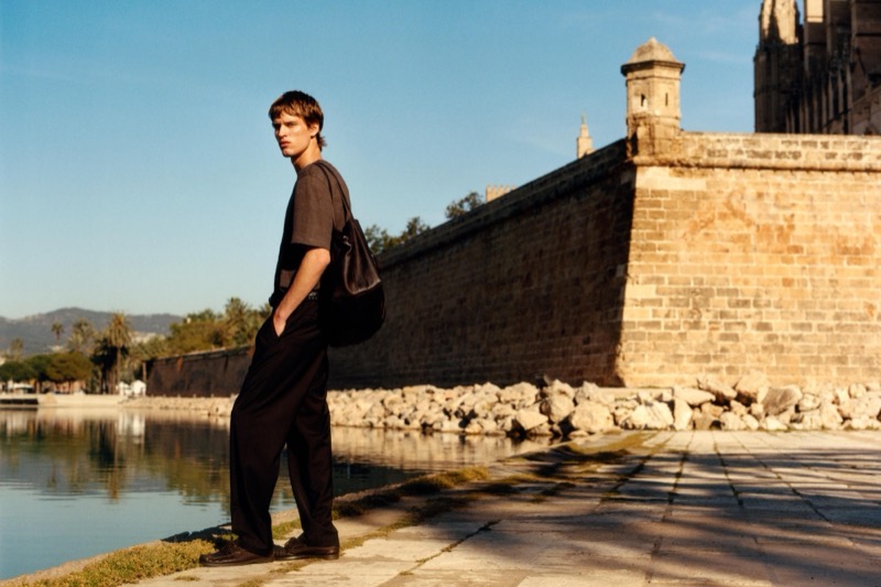 Yeray walks a seaside promenade in a chocolate brown short-sleeve linen tee with matching wide-leg trousers and a brown crossbody bag, the Palma cathedral wall behind him