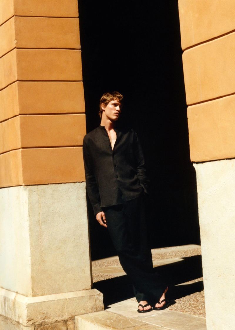 Yeray stands in an ochre stone archway in an unbuttoned black linen stand-collar shirt with matching black trousers and black thong sandals