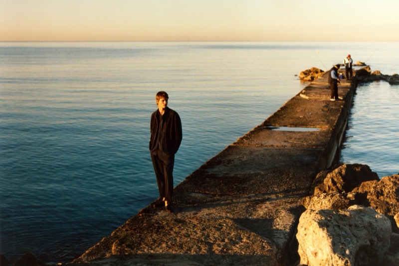 Yeray walks along a stone jetty over the Mediterranean Sea at sunset in an all black linen shirt and trousers
