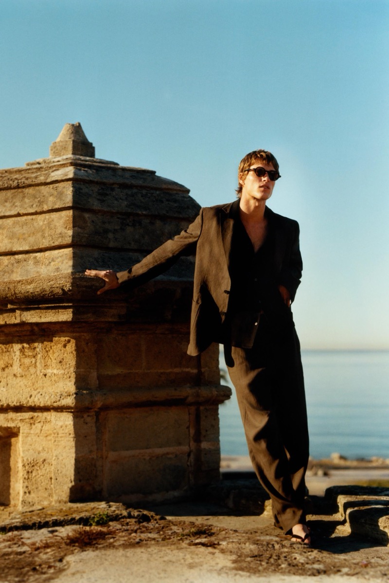 Yeray leans on a stone seaside monument in a black linen blazer over an open black shirt and matching trousers with black sunglasses
