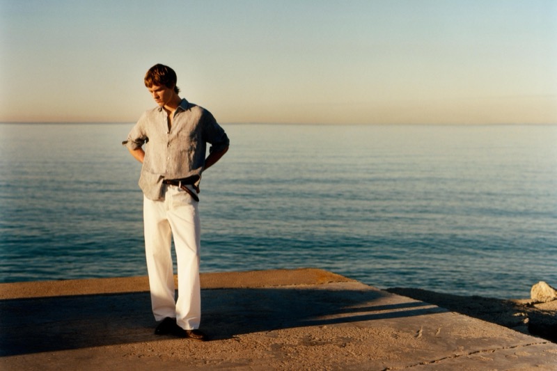 Yeray stands on a stone dock at sunset in a linen shirt with white wide-leg linen trousers