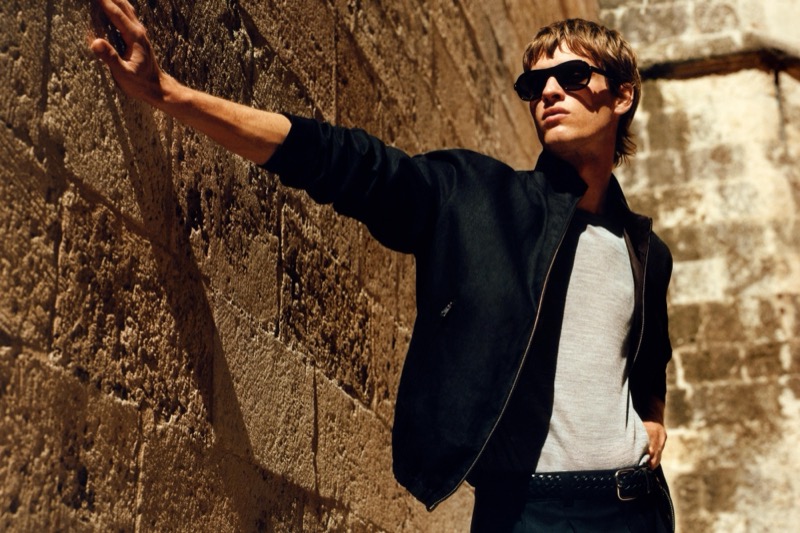 Yeray rests a hand against a sunlit stone wall in a black zip-front jacket over a gray marl tee with black sunglasses and a black studded belt
