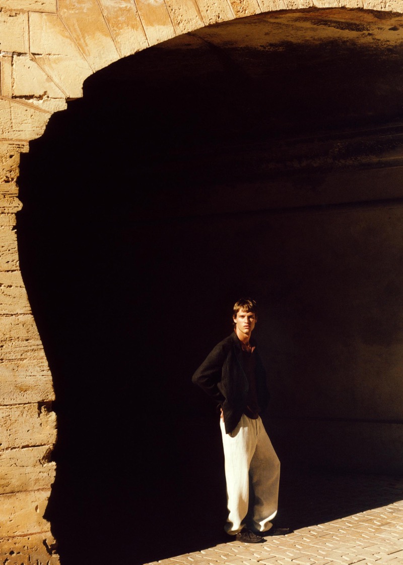 Yeray stands beneath a vast stone arch in a black linen overshirt with white wide-leg linen trousers