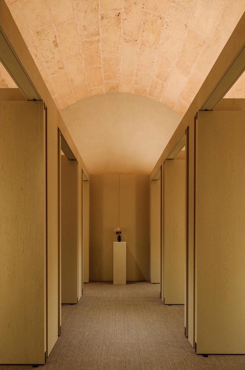 Fitting room corridor at the Palma flagship lined with pale wood doors under a vaulted Mallorcan stone ceiling, ending at a small black vase on a plinth
