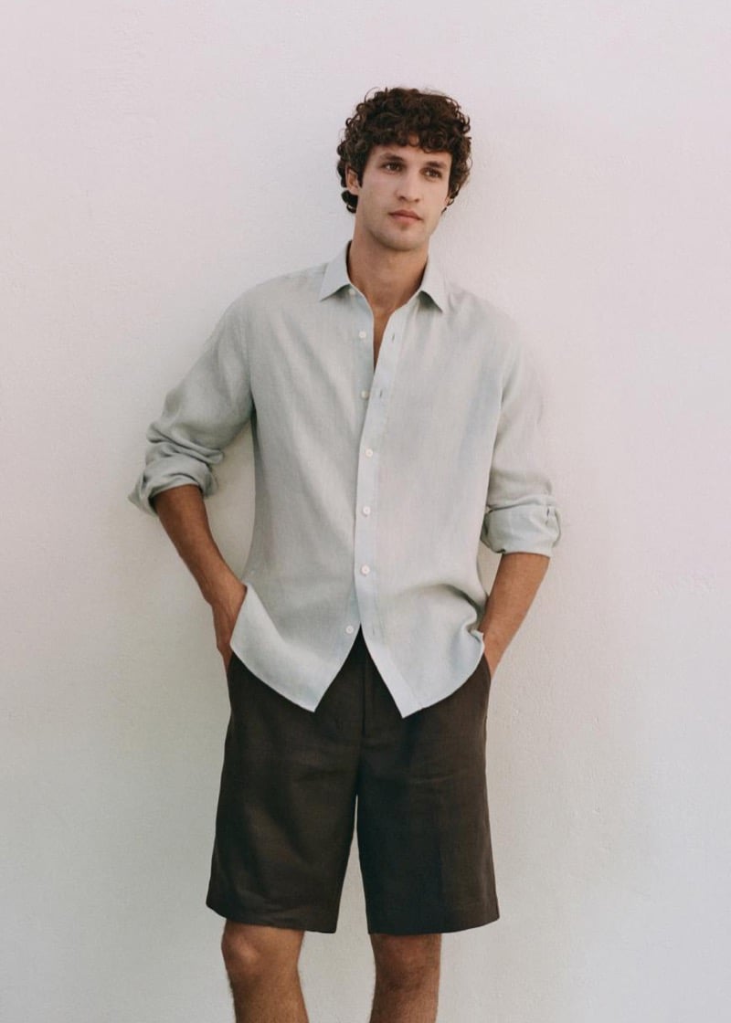 Francisco stands against a pale plaster wall in a linen shirt with rolled sleeves worn half-tucked over dark brown linen Bermuda shorts
