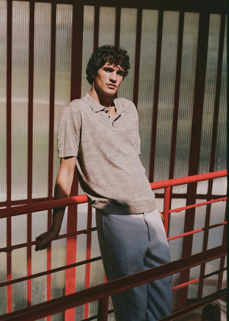 Francisco leans on a red metal railing inside a glass-and-steel atrium in an oatmeal short-sleeve knit polo with a two-button placket and tailored trousers