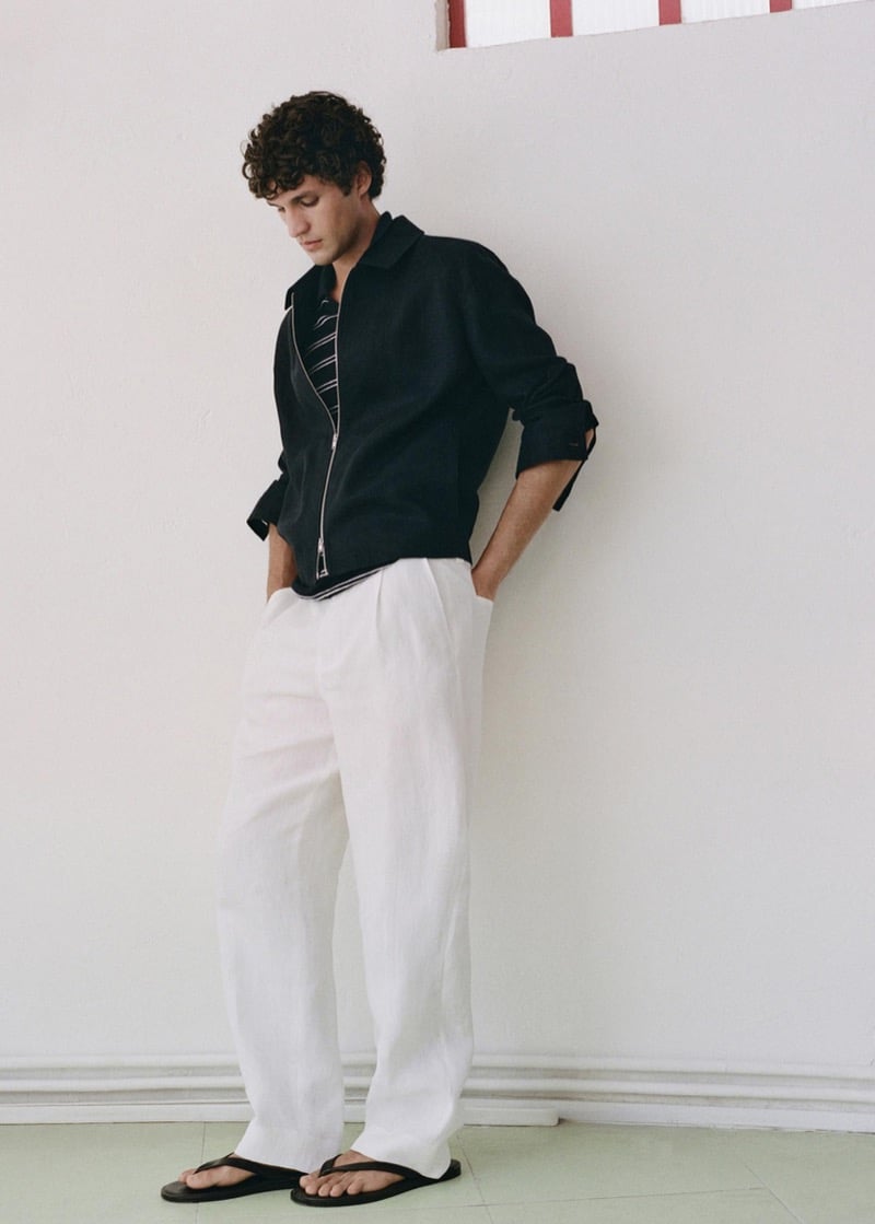 Francisco leans against a whitewashed wall in a black linen zip jacket with strap cuff tabs worn over a black-and-white striped knit polo, paired with white pleated linen trousers and black flip-flops