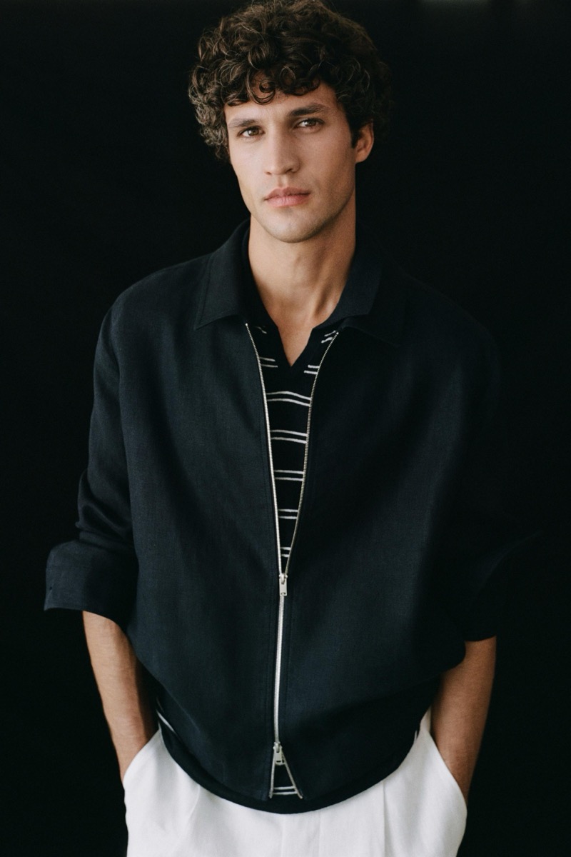 Francisco stands against a dark backdrop in a black linen zip jacket worn open over a black-and-white striped knit polo, white linen trousers cropping at the frame