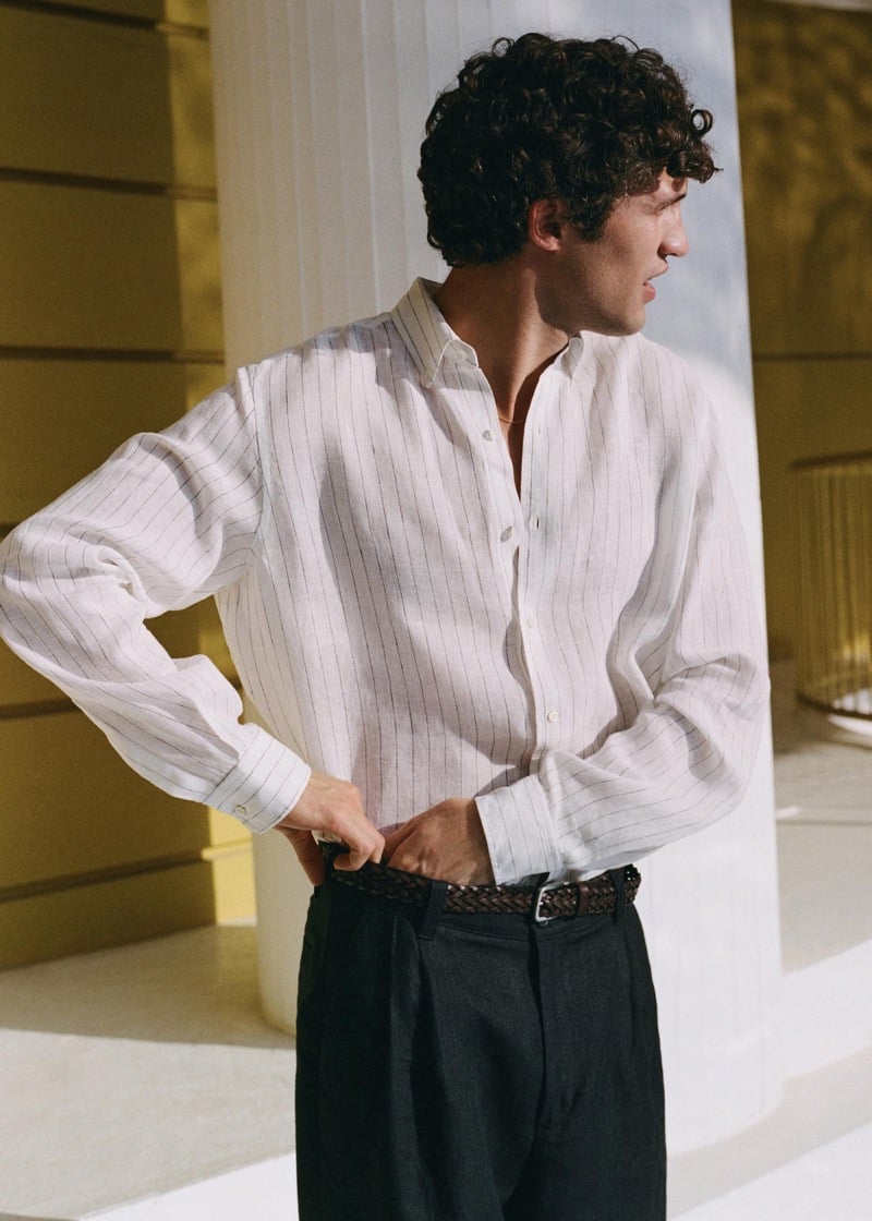 Francisco stands beside a whitewashed column inside a sunlit yellow corridor in a white linen pinstripe shirt with a spread collar, dark pleated trousers, and a braided brown leather belt