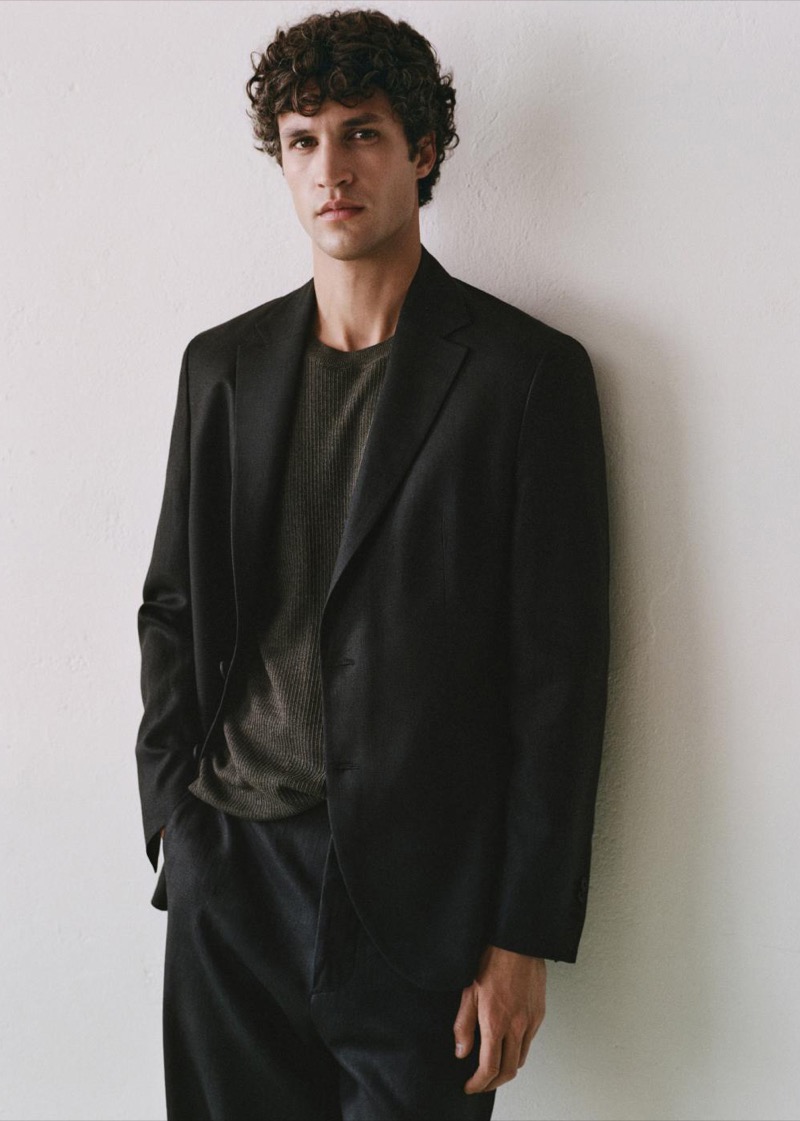 Francisco stands against a pale plaster wall in a soft black single-breasted blazer worn open over a ribbed knit tee and dark pleated trousers