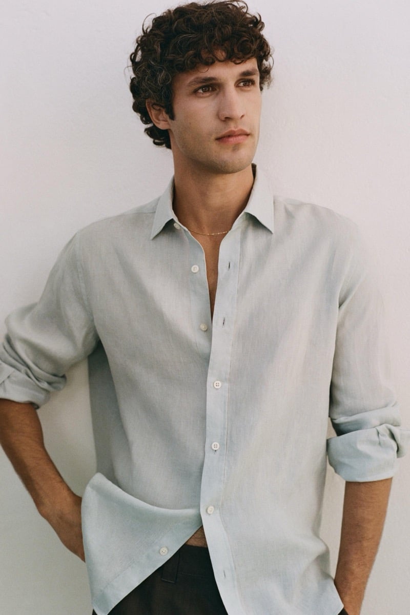 Francisco stands against a pale plaster wall in a pale sage linen shirt with rolled sleeves and the placket open at the chest, hands tucked into the pockets of his trousers