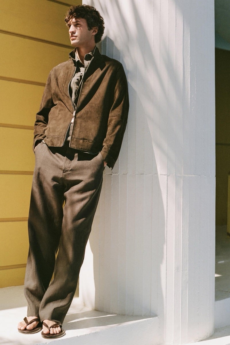 Francisco leans against a whitewashed pilaster beside a yellow building in a brown suede stand-collar zip jacket over a dark olive shirt, taupe linen wide-leg trousers, and brown leather thong sandals