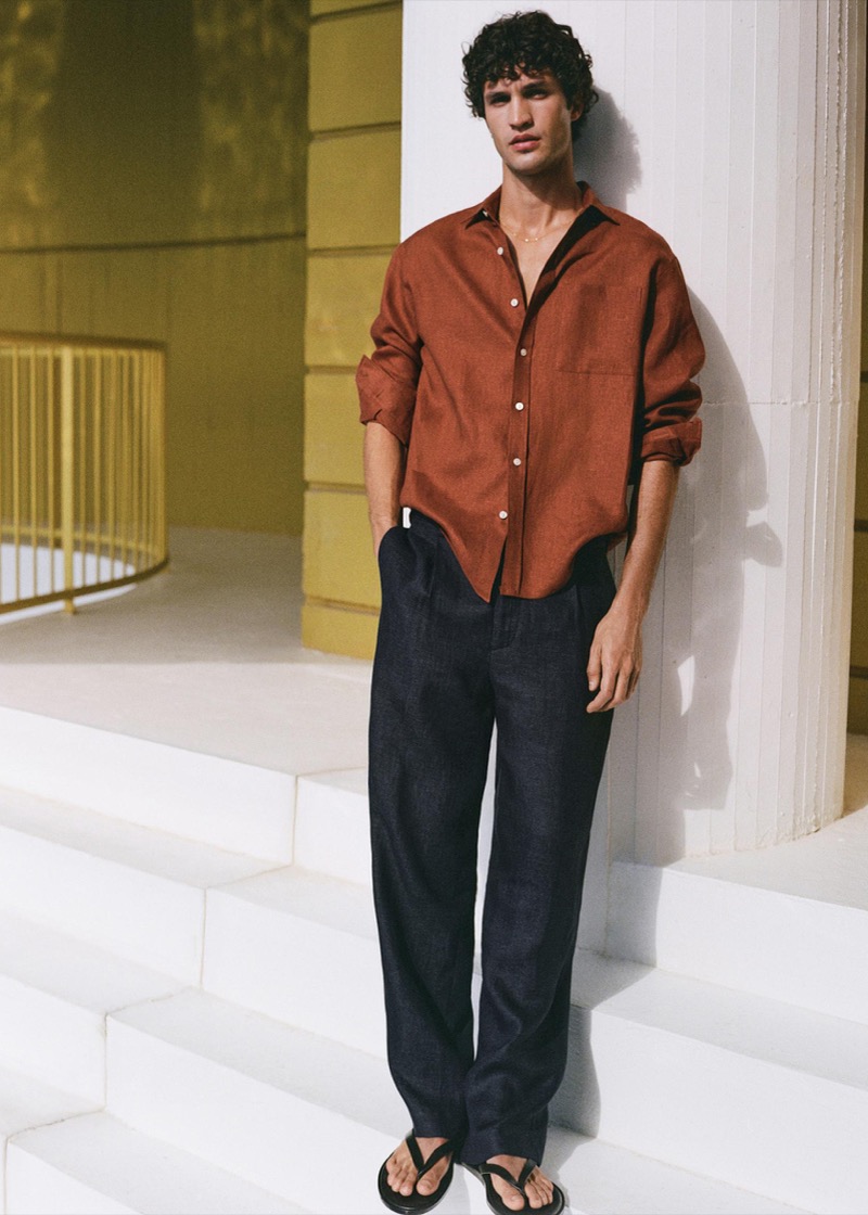 Francisco leans against a whitewashed wall beside a yellow corridor in a rust orange linen shirt with rolled sleeves, dark navy linen wide-leg trousers, and black flip-flops on white stairs