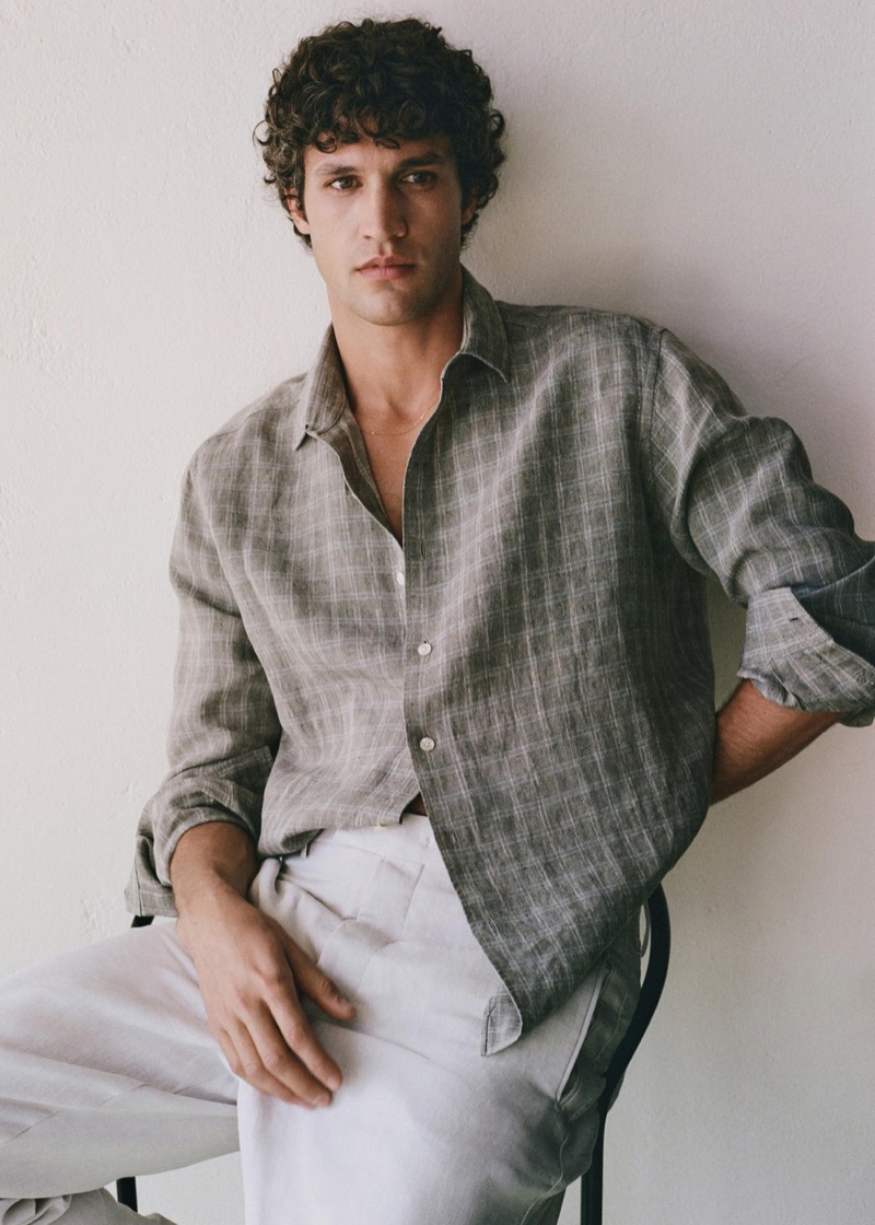Francisco sits on a dark wood stool against a pale plaster wall in an olive gray windowpane linen shirt with rolled sleeves worn over off-white pleated linen trousers