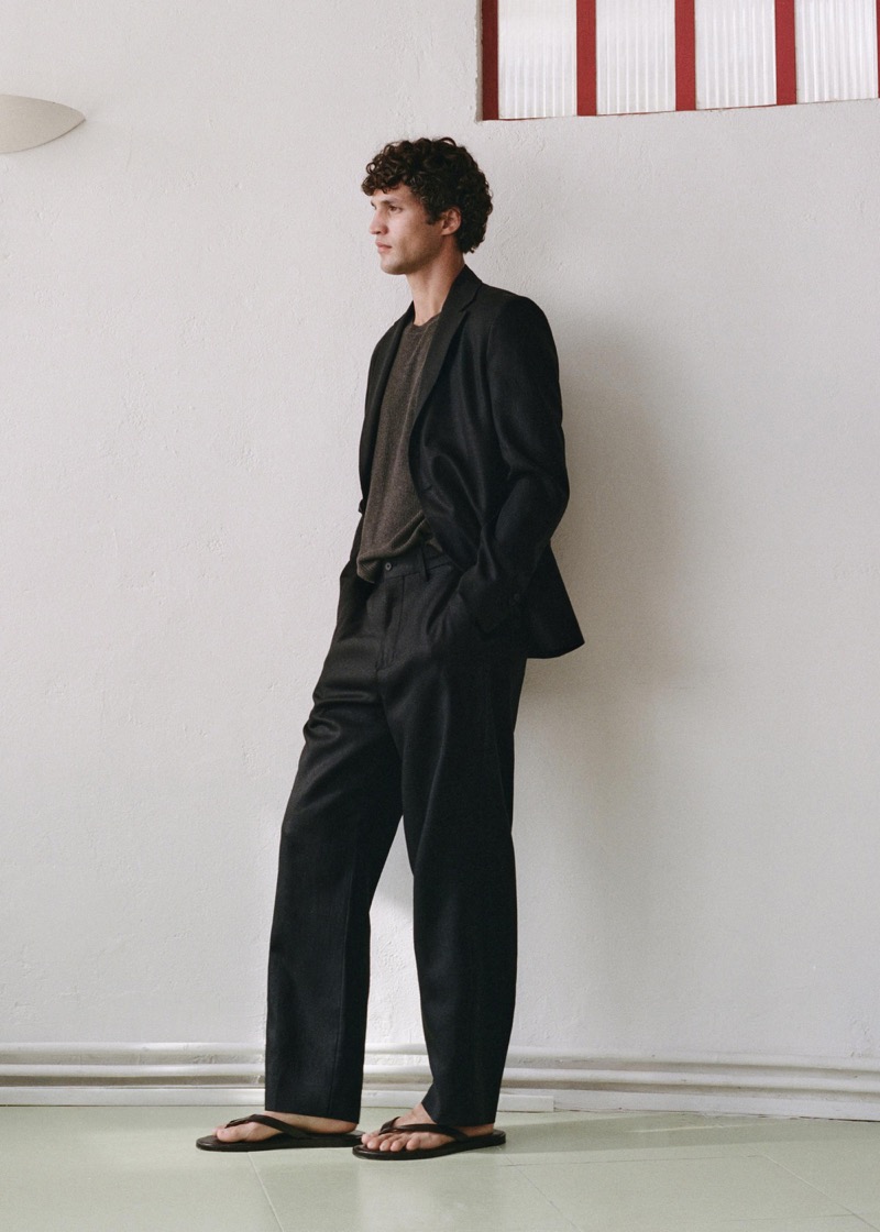 Francisco stands against a pale plaster wall in a black linen single-breasted suit worn over a ribbed knit and finished with black flip-flops
