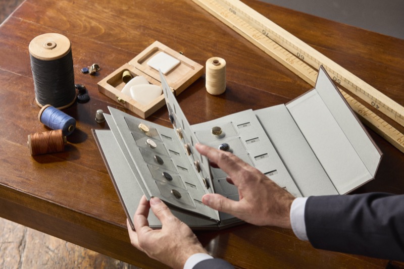 Overhead view of a tailor opening a Canali button presentation book on a polished walnut workbench, surrounded by black, blue, and cognac thread spools, brass thimbles, a small wooden box of chalk and notions, and a yellow boxwood tailoring ruler, the sleeve of a navy suit jacket and a white shirt cuff entering the frame at right