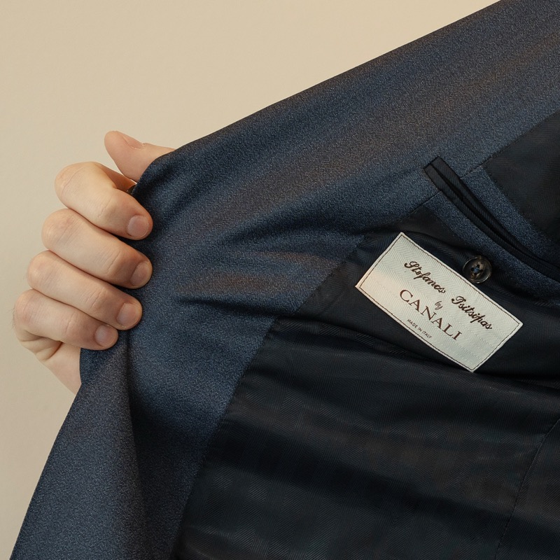 Close-up of the dark blue interior lining of a Canali jacket folded open to reveal a cream silk label embroidered with “Stefanos Tsitsipas by CANALI, Made in Italy,” a hand grasping the lapel from inside the canvas
