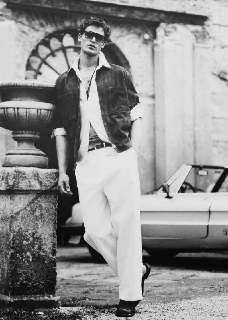 Parker leans against a stone urn in a suede jacket, white shirt, and wide white trousers with sunglasses in a courtyard with a convertible car for Zara