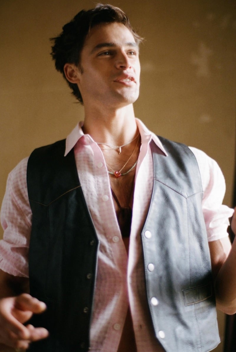 Parker stands in an estate hallway wearing a black leather vest over a pink plaid shirt with white pleated trousers for Zara