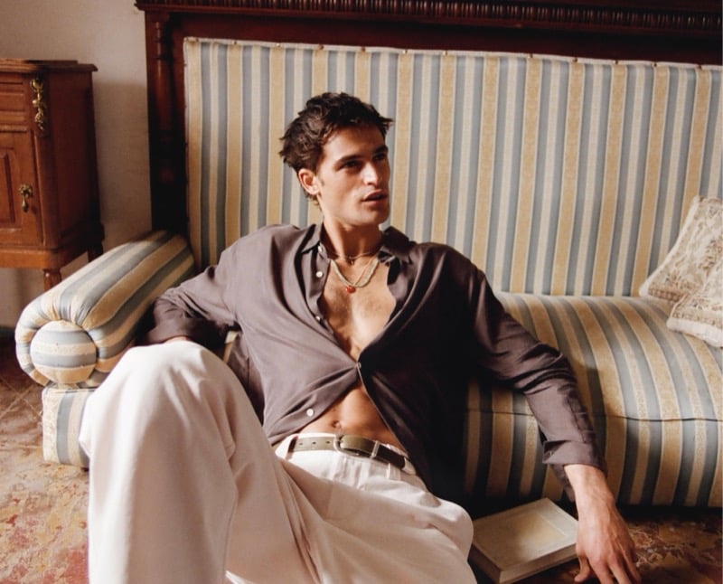 Parker sits on the floor against a striped sofa in an open taupe satin shirt and white trousers with a pendant necklace for Zara