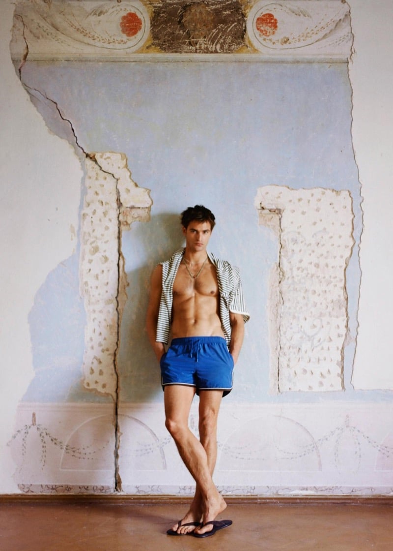 Parker stands in flip-flops and blue swim shorts with a striped hoodie draped over his shoulders against a cracked fresco wall for Zara