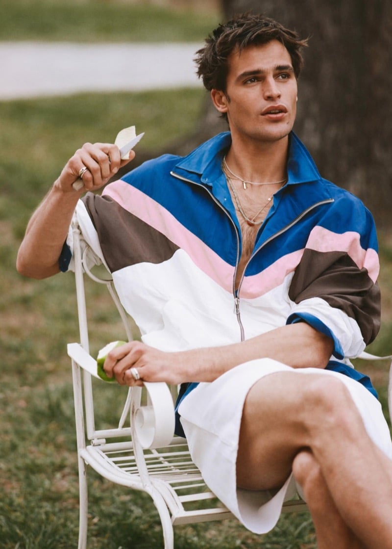 Parker sits in a garden chair wearing a blue, pink, and brown color block jacket with white shorts while peeling an apple for Zara