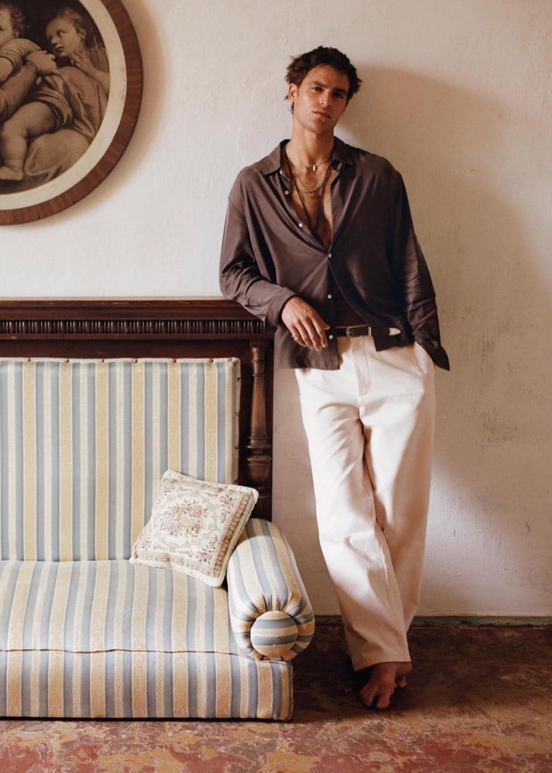 Parker stands barefoot beside a striped sofa in an open taupe satin shirt and wide white trousers with a painting on the wall behind him for Zara