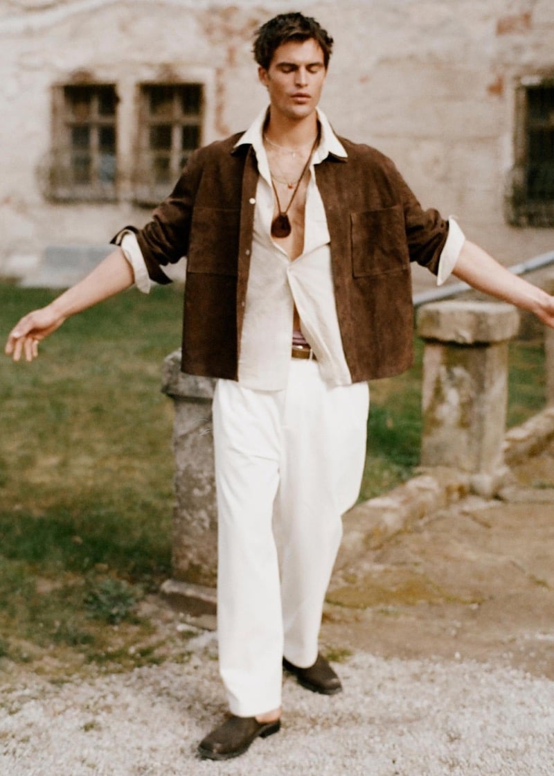 Parker walks across a gravel courtyard in a brown suede overshirt over an off-white shirt and wide white trousers with a pendant necklace for Zara