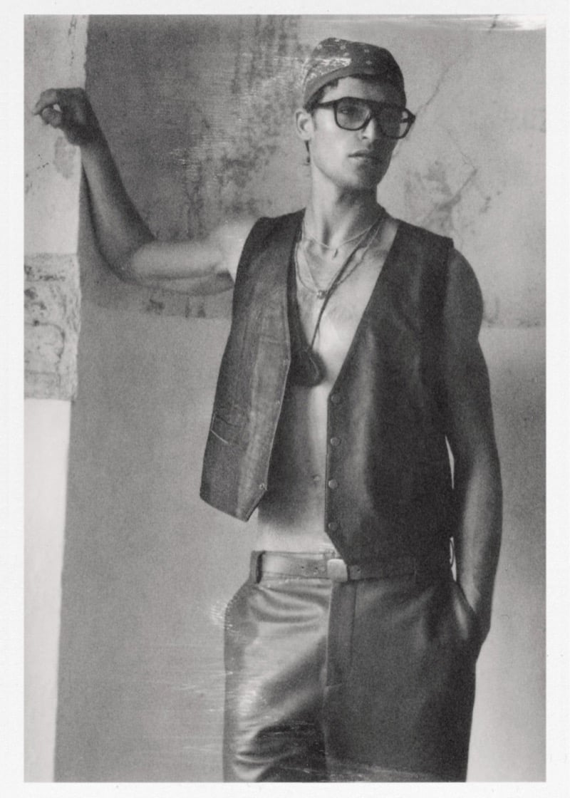 Parker wears a leather vest open on bare skin with a bandana cap and glasses paired with metallic trousers against a cracked wall for Zara
