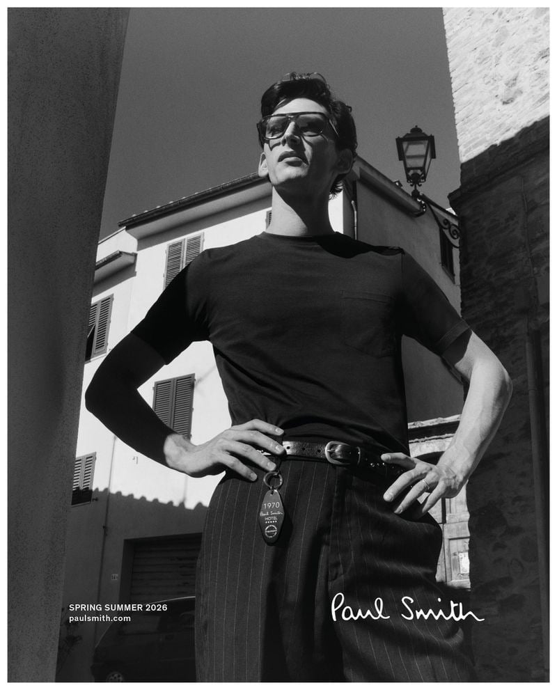 Low-angle black-and-white portrait of Thibaud in a black tee tucked into pinstripe trousers, a Paul Smith Hotel room key dangling from his belt and sunglasses pushed into his hair, framed against an Italian street corner