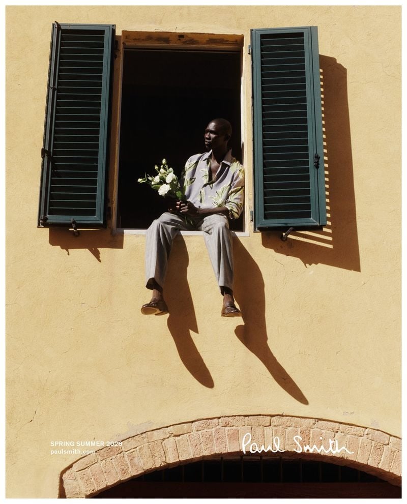 Fernando sits on the ledge of an open window with green shutters set into a yellow Italian stucco facade, holding a small bouquet of white flowers in a printed botanical shirt, gray pleated trousers, and leather sandals