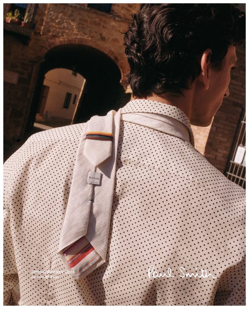 Back-view crop of Thibaud in a cream silk polka-dot shirt with a long Paul Smith striped tie-scarf trimmed in rainbow stripes draped loose down his back, a Tuscan stone archway blurred behind him