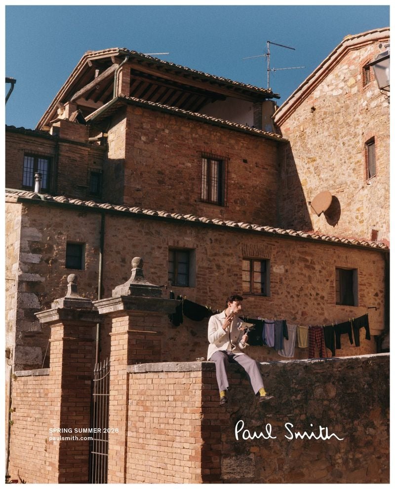 Thibaud perches on a low stone village wall in a cream knit top and gray trousers, a tall Italian stone house and a clothesline of neighbors’ laundry filling the frame behind him