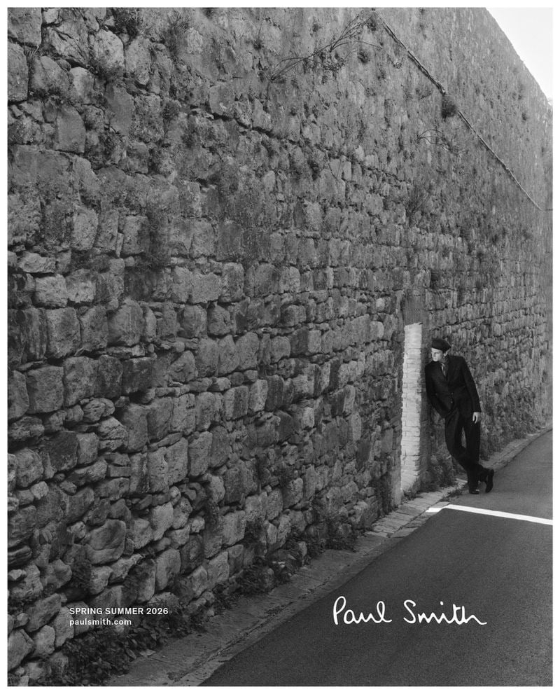 Thibaud leans into a small niche in a tall stone perimeter wall along an empty road, dressed in a dark Paul Smith suit and a flat cap, photographed in wide black and white