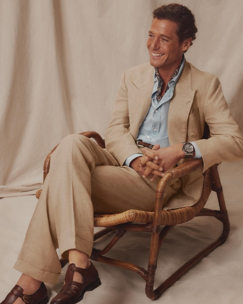 Jon Paul wears a tan peak-lapel linen suit over a light blue shirt with a printed silk neckerchief, seated in a rattan chair with brown leather huaraches