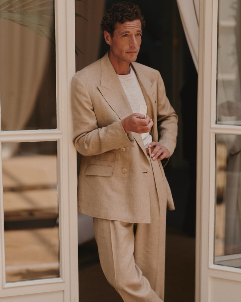 Jon Paul stands in French doors wearing a tan double-breasted peak-lapel linen suit over a white tee
