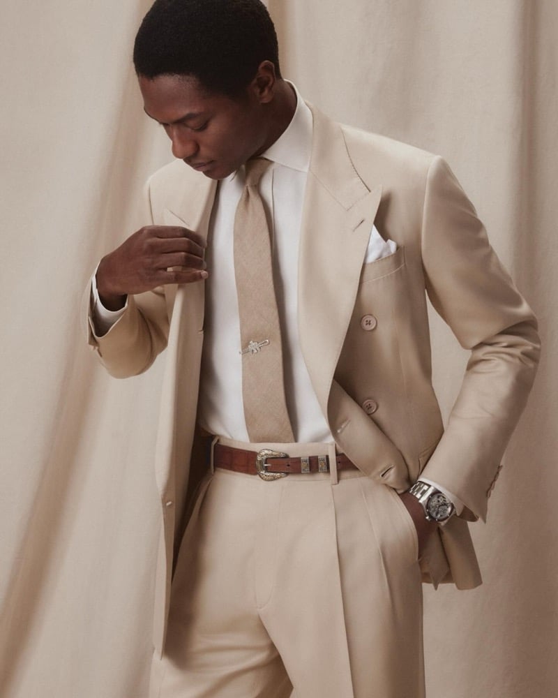Hamid wears a champagne double-breasted peak-lapel suit with a white shirt, tan tie, silver tie clip, and a Western leather belt with engraved silver buckle