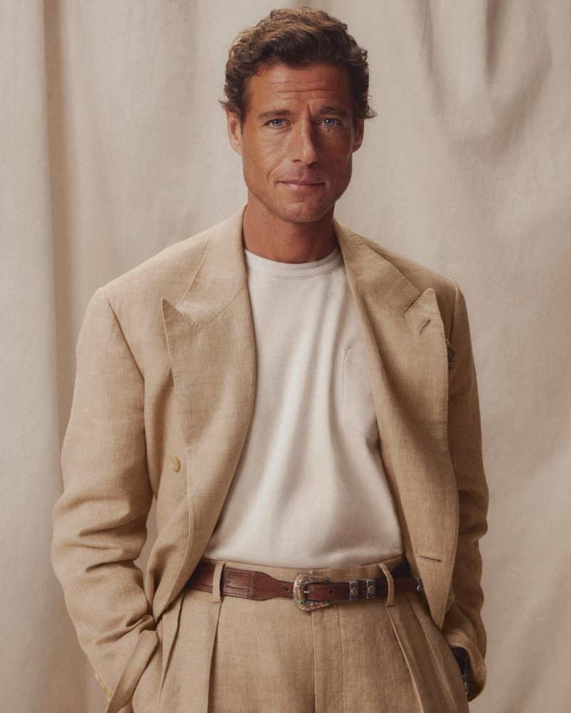 Jon Paul wears a tan double-breasted peak-lapel linen suit open over a white tee with a Western leather belt and engraved silver buckle