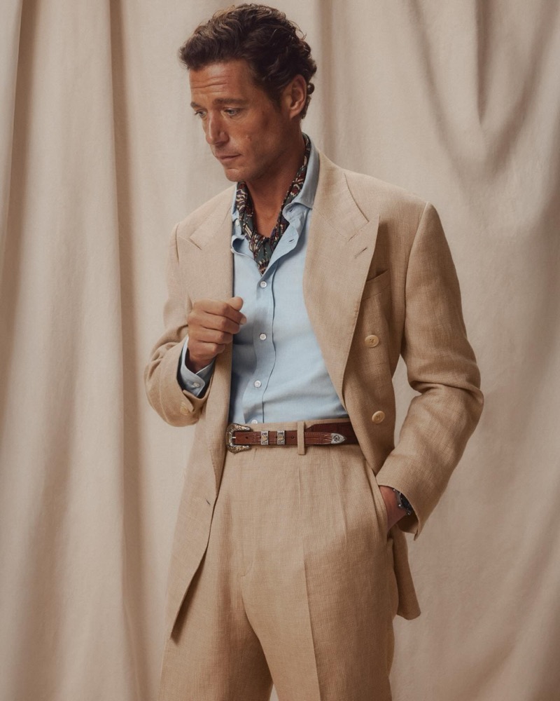 Jon Paul wears a tan double-breasted linen suit with a light blue shirt, printed silk neckerchief, and a Western leather belt with engraved silver buckle