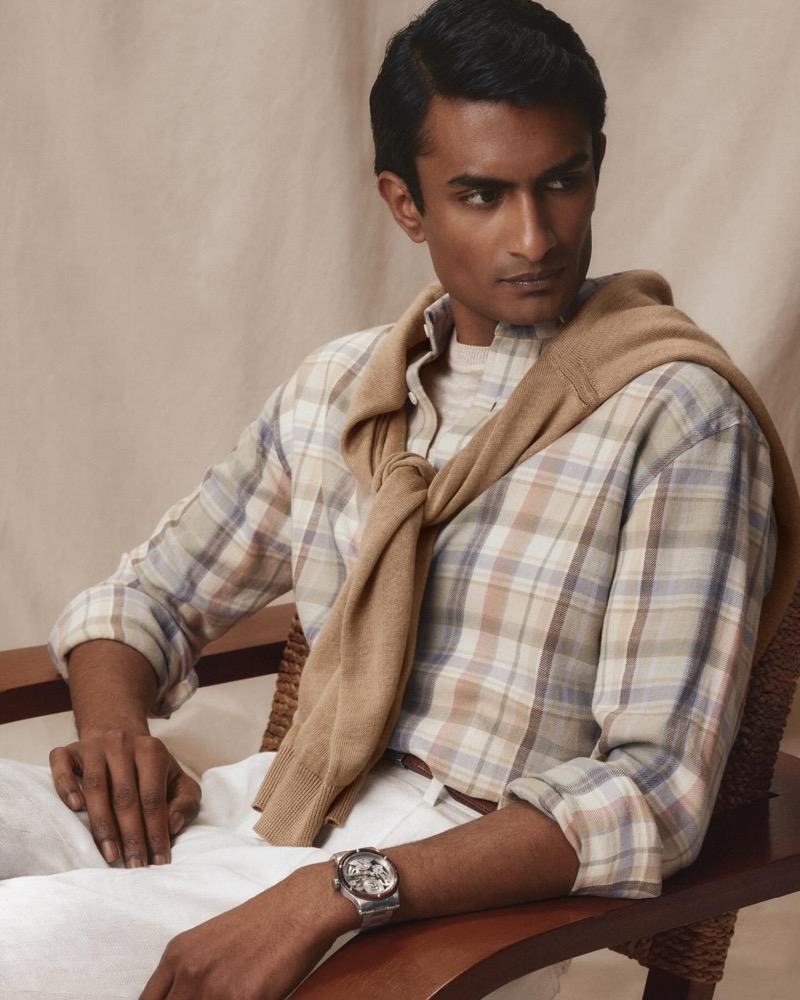 Robin wears a pale plaid linen shirt with a tan crew neck sweater knotted over the shoulders and white trousers
