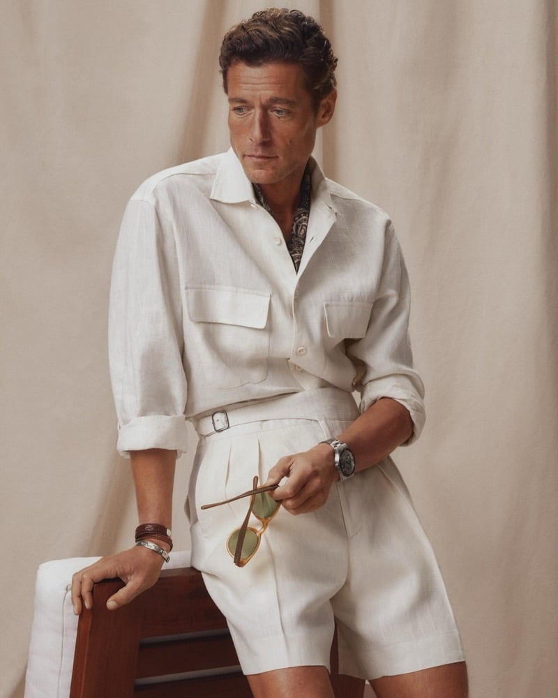 Jon Paul wears a cream linen camp-collar safari shirt tucked into matching pleated shorts with a printed silk neckerchief, holding tortoiseshell sunglasses