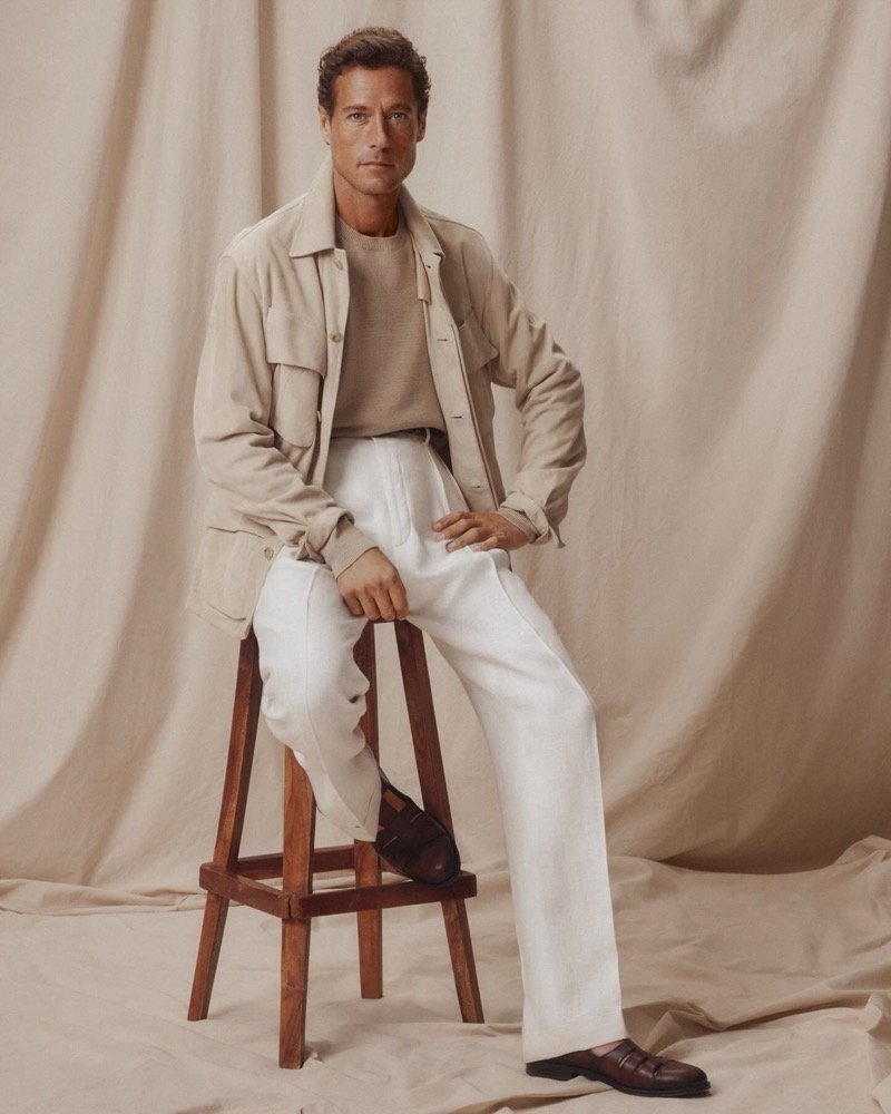 Jon Paul sits on a wooden stool in a tan suede field jacket over a tan crew neck sweater with white trousers and brown leather loafers