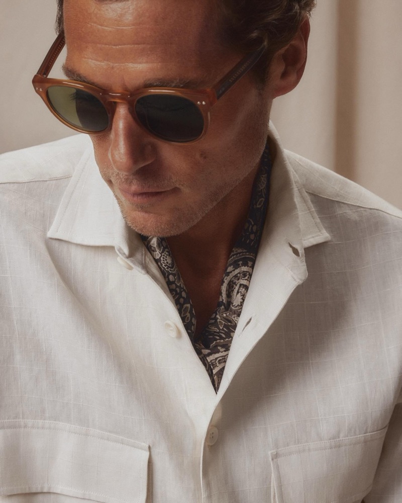 Close-up of Jon Paul in a white linen camp-collar safari shirt with a printed paisley neckerchief and light brown sunglasses