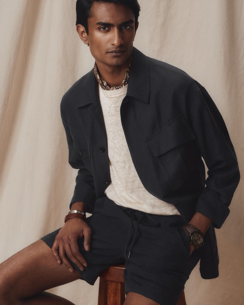 Robin wears a navy field jacket over a cream tee with navy drawstring shorts, a printed neckerchief, and stacked silver and leather bracelets