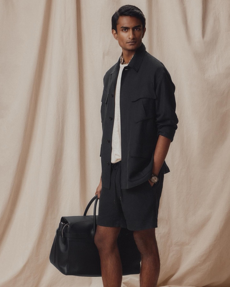Robin wears a navy field jacket over a cream tee with navy drawstring shorts, carrying a black leather bag