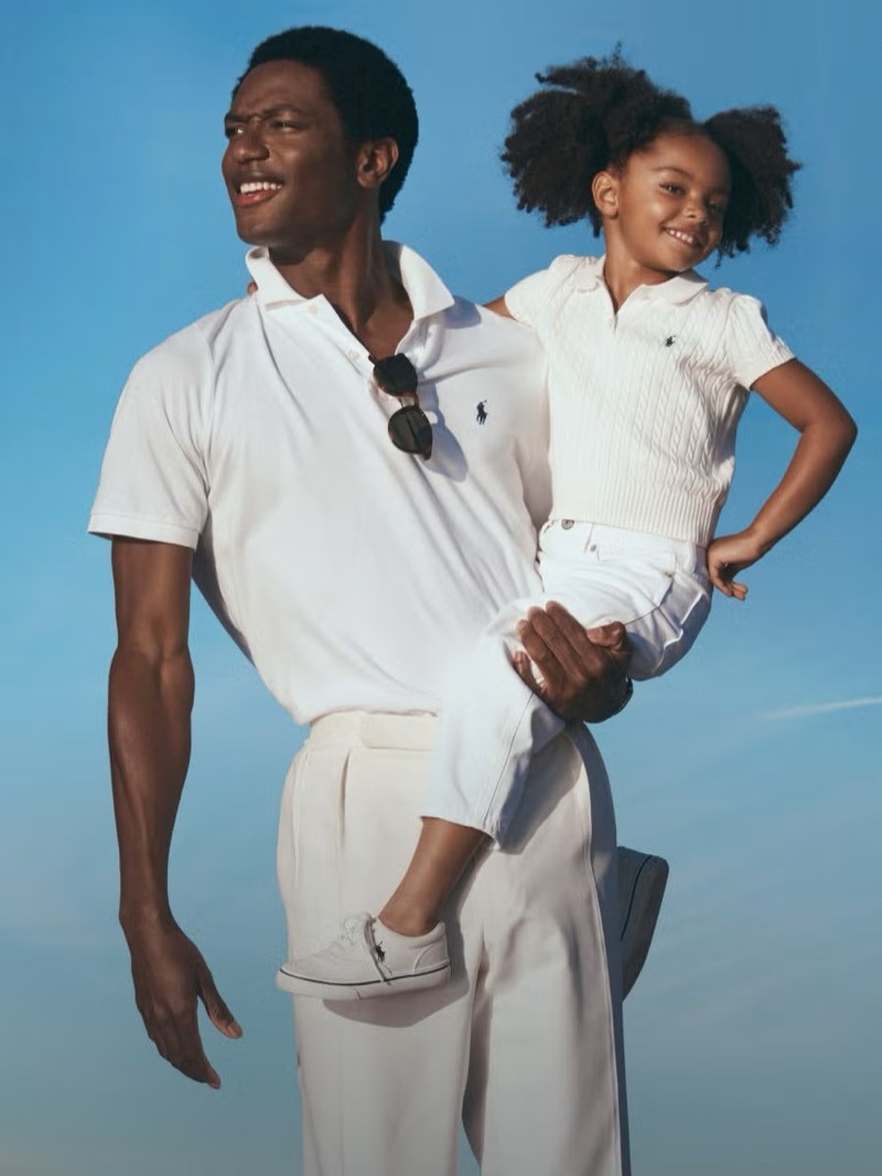 Hamid laughs in a white Polo Ralph Lauren polo shirt with sunglasses hooked at the placket, pleated white trousers, and white Polo sneakers, lifting a small girl in a white cable-knit polo and matching trousers onto his hip against an open sky