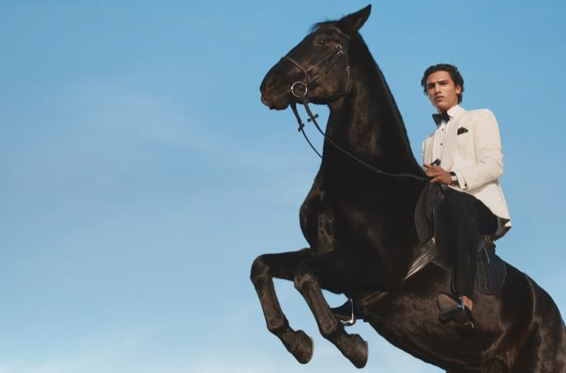 Morgan rides a black horse rearing against a clear blue sky in a white Purple Label tuxedo dinner jacket with a black bow tie, black trousers, and black evening loafers