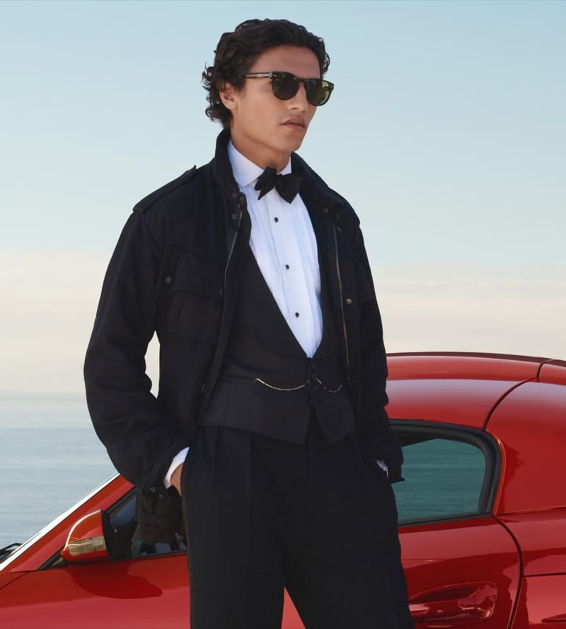 Morgan leans against a red sports car at the coast in dark sunglasses, a black field jacket open over a white tuxedo shirt with a black bow tie and black tuxedo waistcoat, and black tuxedo trousers, the sea behind him